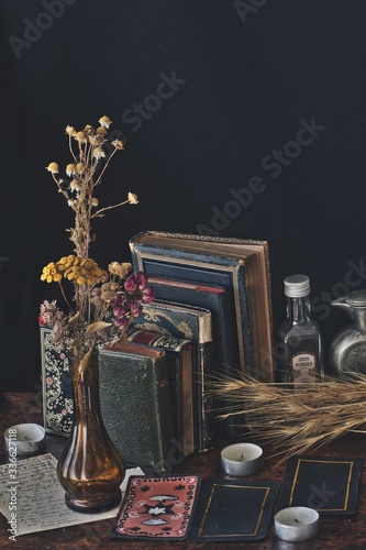 Messy wiccan witch altar filled with old vintage books, dried chamomile and rose flowers in rustic bottles, herbs, white candles, hand painted tarot cards, postcards in black background. Vertical
