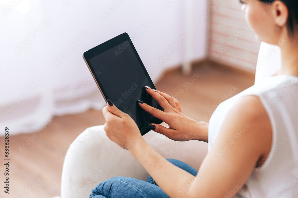 Naklejka premium A young girl sits in chair with tablet and pays bills, online purchases, payment for goods during quarantine, A young girl undergoes online training, online training during quarantine, work from home