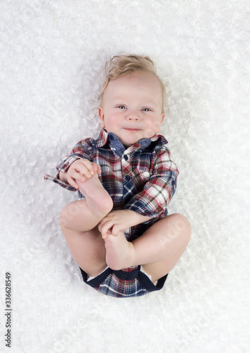 a six-month-old blond boy with blue eyes in a checkered shirt bodik lies on his back playing with his legs, smiling cute, on a white background