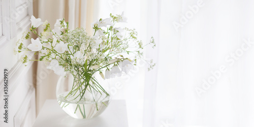 Bouquet of gentle bells in vase. Morning light in the room. Soft home decor, glass vase with white flowers on  white wall background and on wooden table. Interior. Greeting card. Copy space. Banner
