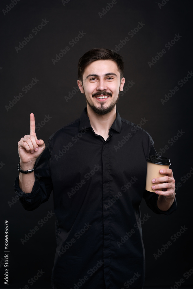 Portrait of smiling handsome young man with mustache and beard holding coffee cup and raising index finger up while having idea Portrait of smiling handsome young man with mustache and beard holding coffee cup and raising index finger up while having idea