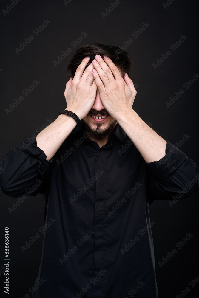 Smiling young man with beard covering eyes with hands in anticipation of surprise against black background Smiling young man with beard covering eyes with hands in anticipation of surprise against black background