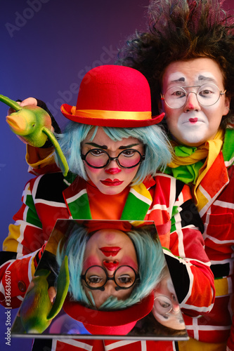 two clowns a man and a woman with bright makeup in colored costumes say they look at their reflection in the mirror. 