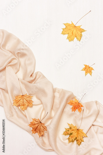 Autumn cozy composition with dried leaves of maple and pastel beige scarf on white wooden background. Autumn, fall concept. Flat lay, top view, copy space.