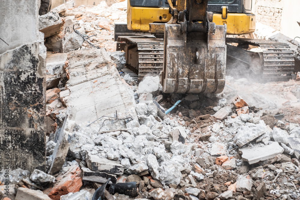 Fototapeta premium Small yellow excavator is on duty for pulling down the old buildings. Selective focus at bucket.