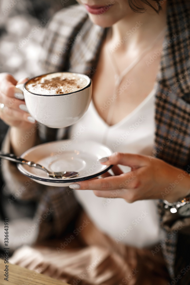 Close-up of a white cup of coffee and a white saucer in the hands of a beautiful girl of European race. She's wearing a checkered jacket.