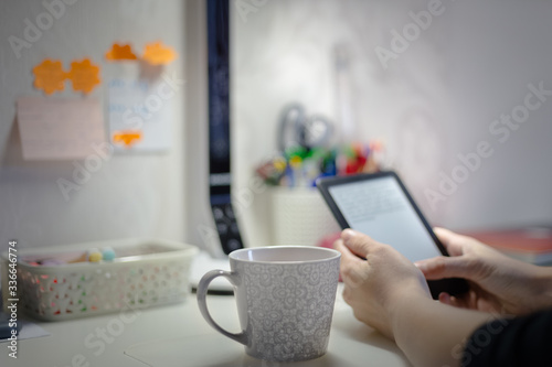 Young woman using tablet , reading a book at home at her desk and drinking some hot beverage