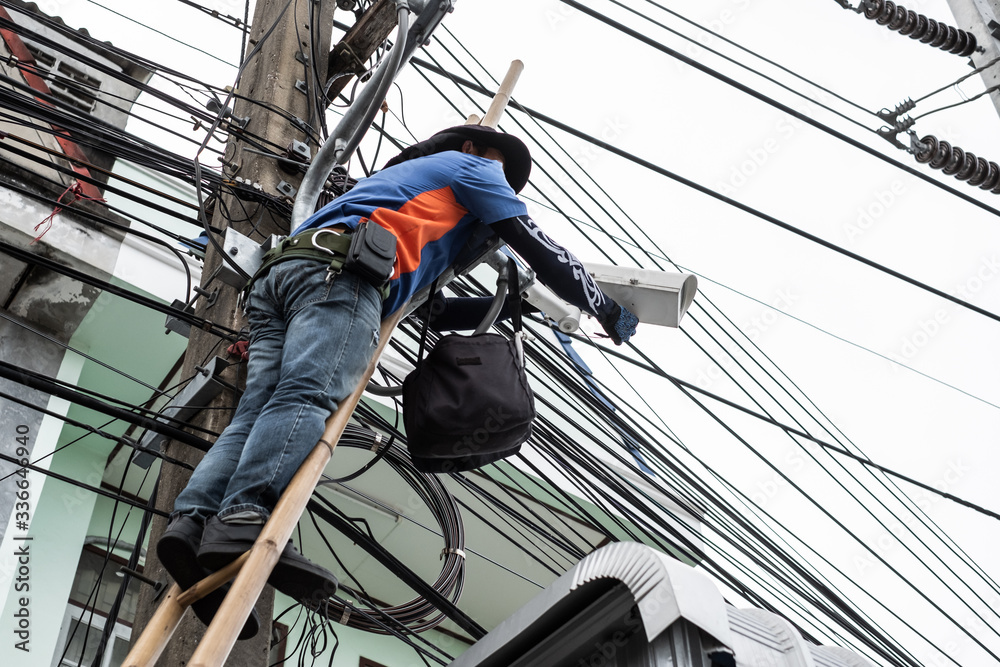 Technician worker climbing wooden bamboo to instal CCTV camera security system on public electricity pole.