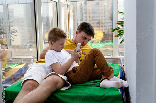 A young father and little son are lying on a mattress on a loggia. Self-isolation during a pandemic COVID-19. A little boy looks at his cell phone, a young man uses the Internet on his cell phone. The