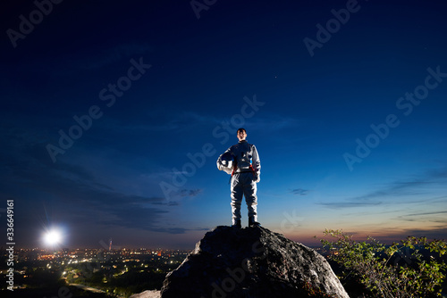 Brave astronaut standing on top of rocky hill with beautiful blue sky, city lights on background. Handsome spaceman in white space suit holding helmet and looking at camera. Concept space travel.