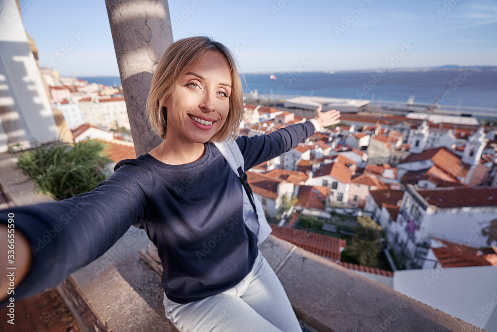 Traveling by Portugal. Young traveling woman taking selfie in old town Lisbon with view on red tiled roofs, ancient architecture and river.