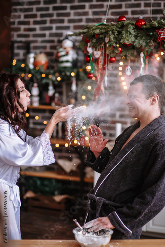 A guy and a girl in dressing gowns in the kitchen  which is decorated for the celebration of Christmas and New year. Guys play with flour