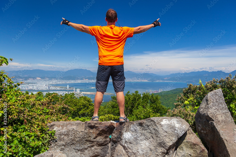 a male backpacker  on top of a mountain looks at Danang bay in a traditional Vietnamese hat