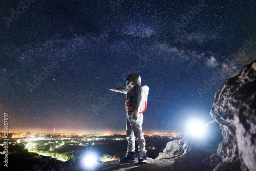 Male spaceman pointing finger at night city in valley while standing on rocky mountain under fantastic night sky with stars and Milky way. Concept of cosmonautics and space traveling.