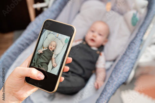 Mother takes a picture with her son by phone. The woman will send photos to grandparents via email. Mom is spending time with her cute baby boy at home office. Mother's love. Maternity leave content. photo