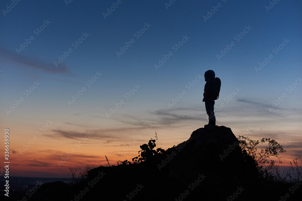 Silhouette of astronaut standing on top of rocky hill with beautiful night sky on background. Space traveler wearing space suit with helmet. Concept of space travel.