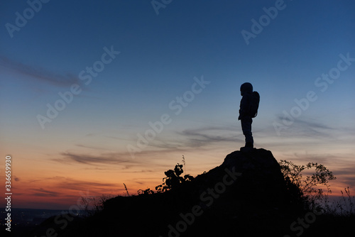 Silhouette of astronaut standing on top of rocky hill with beautiful night sky on background. Space traveler wearing space suit with helmet. Concept of space travel.