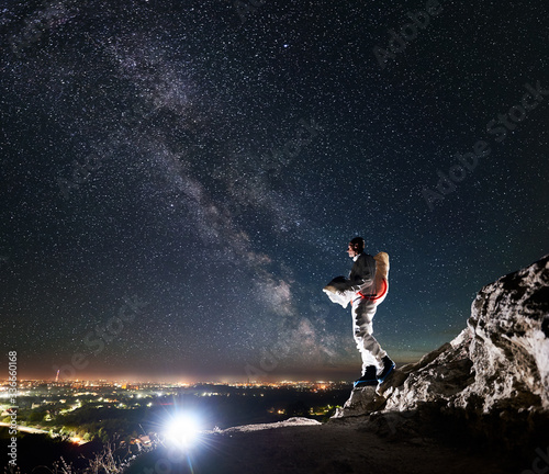 Cosmonaut standing on rocky hill under beautiful night sky with stars and Milky way. Space traveler in space suit holding helmet and looking at night city lights. Concept of cosmonautics and nighttime