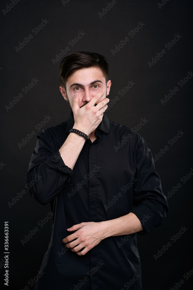 Portrait of shocked young brunette man in black shirt covering mouth with hand, not believing his eyes Portrait of shocked young brunette man in black shirt covering mouth with hand, not believing his eyes