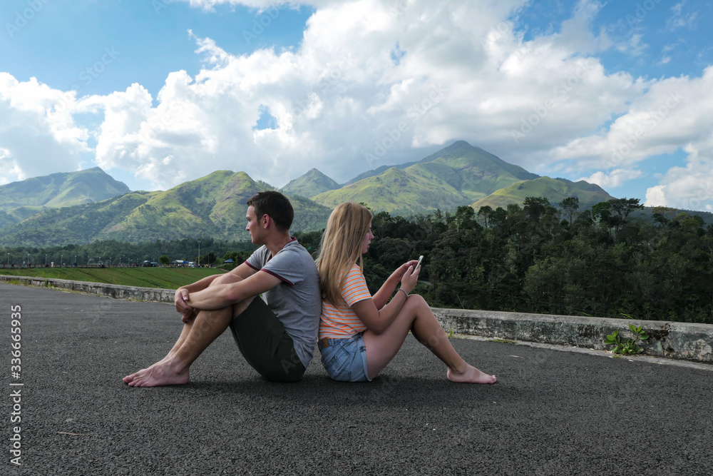 Girl and boy sitting on the road to the mountains. Green mountains with blue sky and clouds. Couple in the nature.