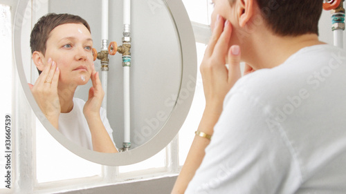 Caucasian woman with a short haircut in front of mirror in bathroom full of sunlight put soma face cream on and massage photo