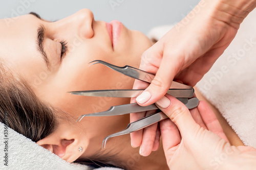 Hands of cosmetologist holds tweezers for eyelash extensions on background of face of young woman  close up.