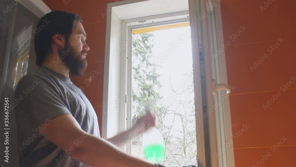 A man in a gray t-shirt and a beard uses cleaning agent and a sponge to wash the Windows of the house