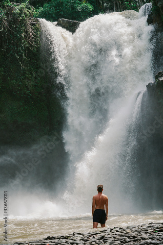 A young man standing under a big / tall waterfall in Indonesia.