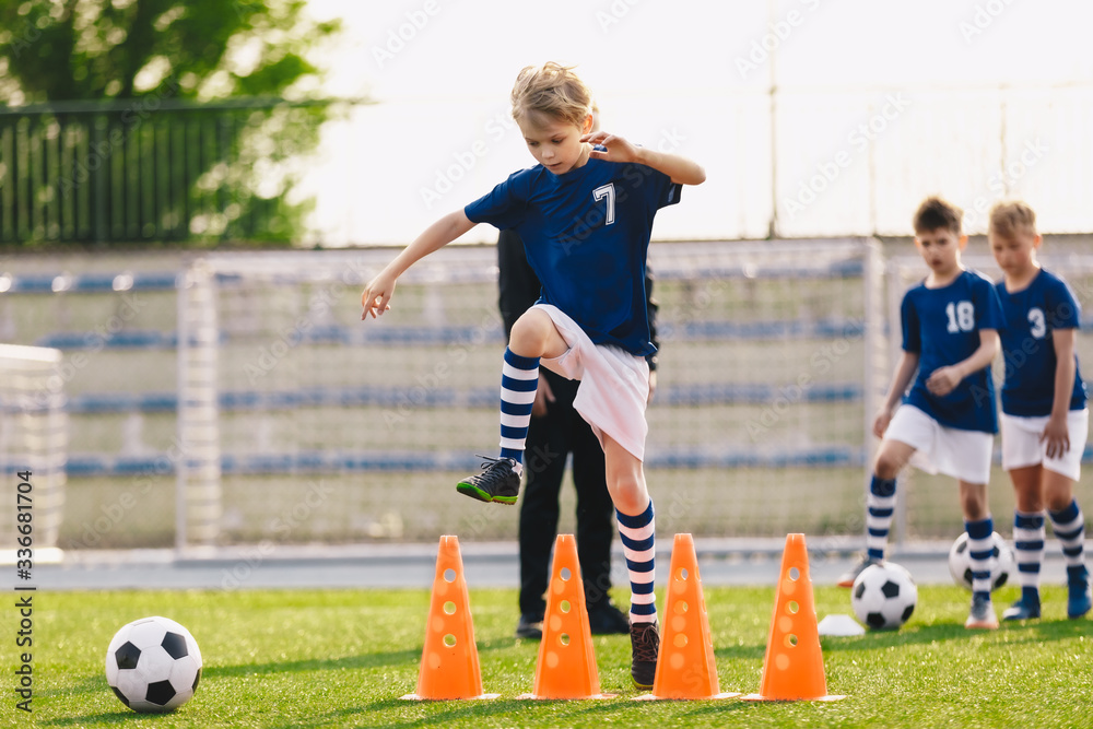 Obraz premium Boy jumping over soccer cones on football training field. Kids on sports training with junior coach. Children on physical education class. Boys imporoving soccer skills