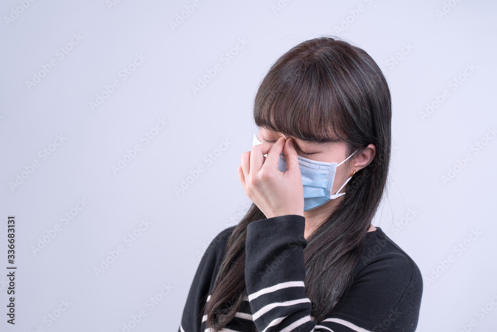 Young woman is feeling dizzy, headache, sick, unwell with wearing a medical blue face mask over the gray background, close up, copy space. Young woman is feeling dizzy, headache, sick, unwell with wearing a medical blue face mask over the gray background, close up, copy space.