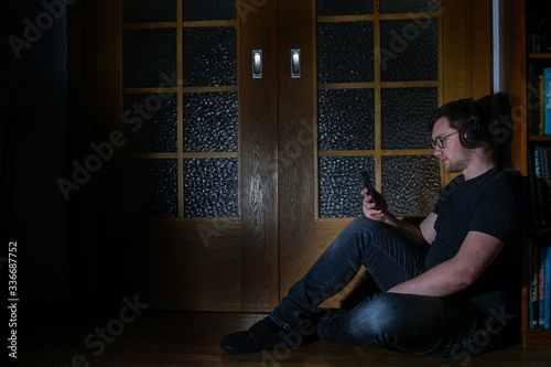 Mann in schwarzem T-Shirt sitzt auf dem Boden und schaut auf sein Smartphone. Mann sitzt in einem dunklen Raum auf dem Boden vor einer Holztür. Low Key Fotografie.