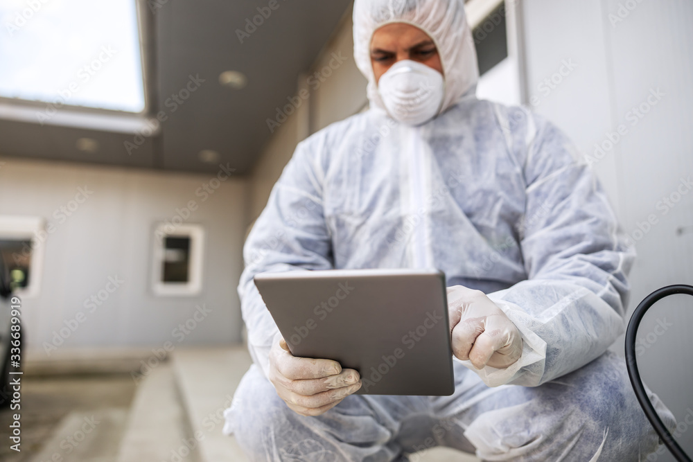 Man in virus protective suit and mask looking and typing on tablet, disinfecting buildings of coronavirus with the sprayer. Infection prevention and control of epidemic. World pandemic. Man in virus protective suit and mask looking and typing on tablet, disinfecting buildings of coronavirus with the sprayer. Infection prevention and control of epidemic. World pandemic.