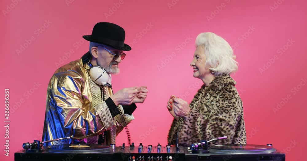 Sylish senior man in golden sparkling jacket and hat and woman with gray hair in leopard coat having fun and DJing on pink background. Couple of old djs dancing playing music on mixer. Disco concept.