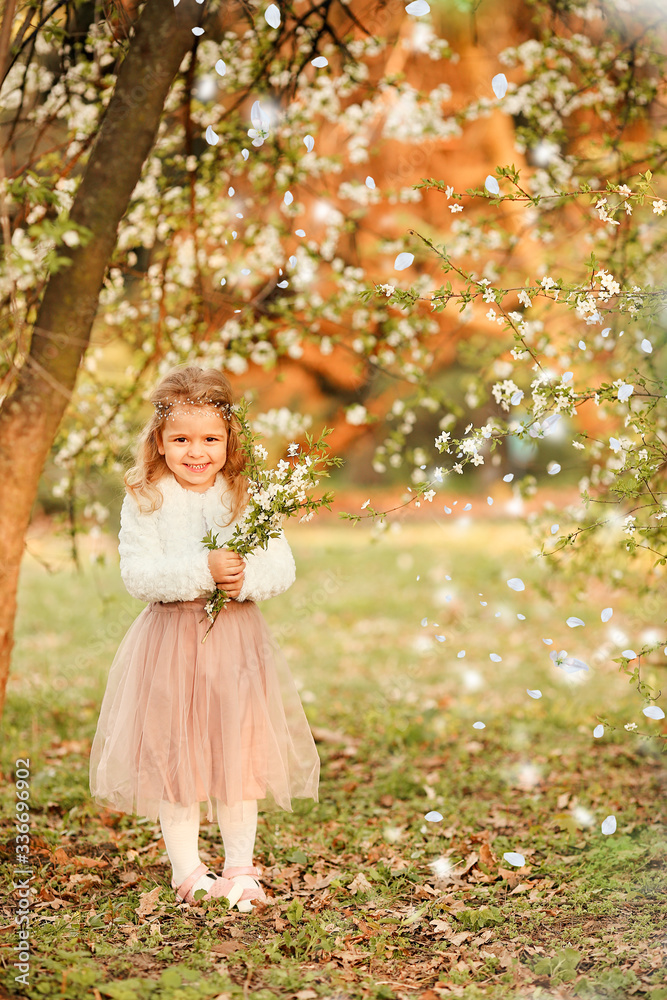 A beautiful cute little girl laughs and has fun in a flying nude skirt and a white fur blouse with a bouquet of blossoming cherry flowers in a spring botanical garden. For Easter and spring cards A beautiful cute little girl laughs and has fun in a flying nude skirt and a white fur blouse with a bouquet of blossoming cherry flowers in a spring botanical garden. For Easter and spring cards