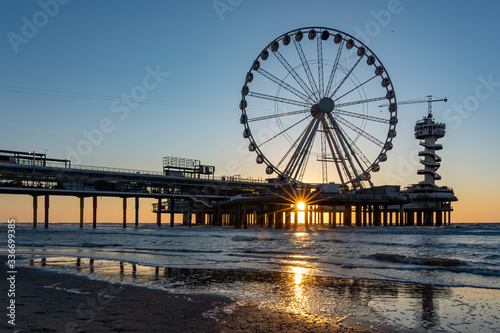 The setting sun is just below the pier in Scheveningen with the ferris wheel and the bungy jump tower, and glistens on the wet sand on the water line of the North Sea