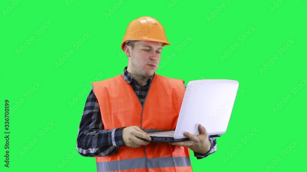 A man in a construction helmet and an orange vest is talking on a laptop in a good mood, green screen background A man in a construction helmet and an orange vest is talking on a laptop in a good mood, green screen background