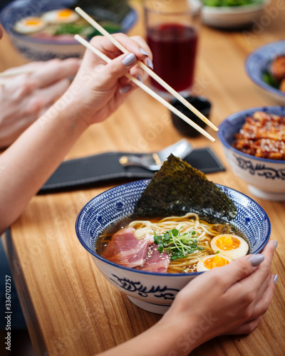 Female with chopsticks eating asian japanese shio ramen noodle soup with eggs, pastrami and spinach, vertical photo