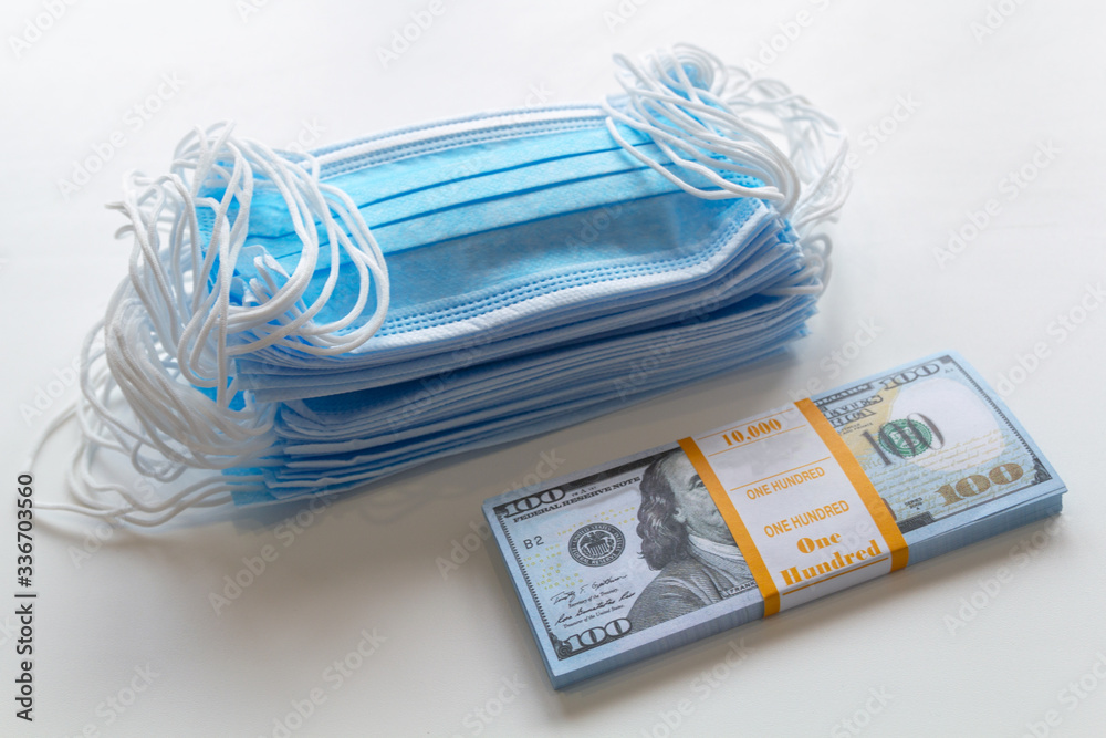 A pack of medical masks and a stack of 100 dollars bills as a symbol of higher prices and a deficiency in the protection of the respiratory tract from viruses. A pack of medical masks and a stack of 100 dollars bills as a symbol of higher prices and a deficiency in the protection of the respiratory tract from viruses.