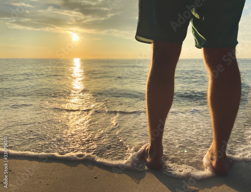 Man in shorts meets beautiful sunset, sunrise.Warm sea wave with foam touches the legs. Sandy beach and tranquil place on the sea