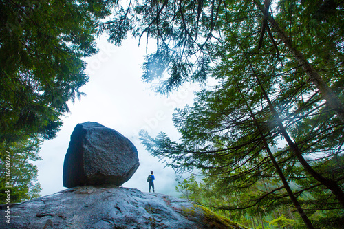 Woman hiker stands next to a huge boulder surrounded by trees of the foggy forest in Squamish on the Stawamus Chief hike.