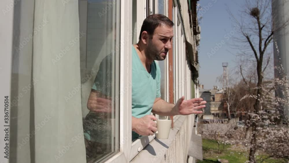 A disgruntled man stands near an open window with a cup in his hand, shows signs of disagreement and denial, leaves and closes the window. camera movement through the window frame, from left to right A disgruntled man stands near an open window with a cup in his hand, shows signs of disagreement and denial, leaves and closes the window. camera movement through the window frame, from left to right