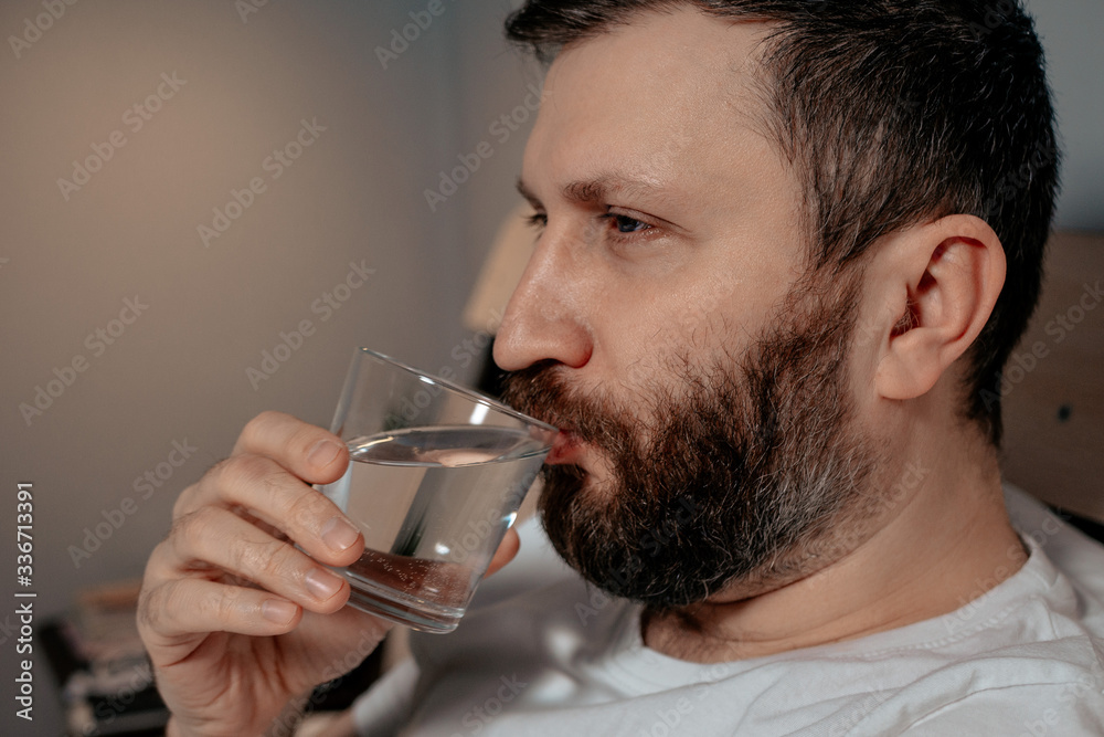 A lonely bearded dark-haired man drinks water from a glass while lying on his bed in the evening. Close-up on the right, healthy habit, bedroom, self-isolation. A lonely bearded dark-haired man drinks water from a glass while lying on his bed in the evening. Close-up on the right, healthy habit, bedroom, self-isolation.
