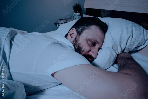 A lonely bearded dark-haired man sleeps soundly lying on his stomach at night on the bed, hands clasped. Close-up on the right, white bedding, bedroom, self-isolation.