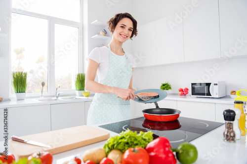 Photo of beautiful cheerful housewife lady putting fresh raw salmon fillet steak on flying pan keeping diet morning cooking wear apron t-shirt stand modern kitchen indoors