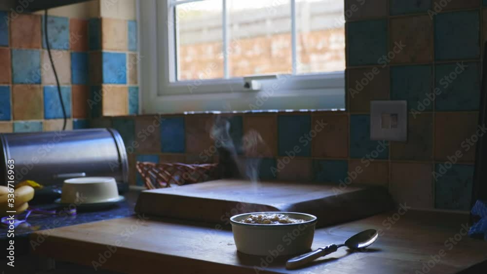 Steaming bowl of porridge and spoon on wooden countertop in old cottage kitchen. Near window and coloured tiles on wall. Metal bread bin and bananas on the side. Steaming bowl of porridge and spoon on wooden countertop in old cottage kitchen. Near window and coloured tiles on wall. Metal bread bin and bananas on the side.