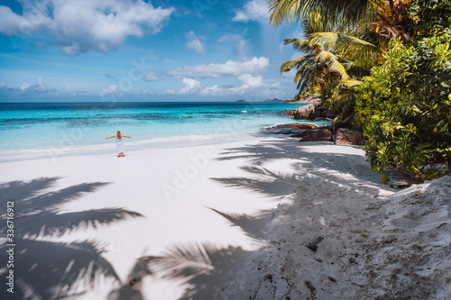 Female tourist with raised hands enjoy empty tropical beach on vacation. White powdery sand beach, palm trees and blue ocean lagoon. Exotic paradise recreation vacation concept