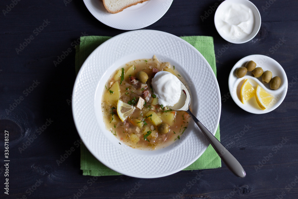 Flat lay on Solyanka soup with fermented cabbage, potato, meat, lemon, olives, sour cream, bread, spoon  on black wooden background