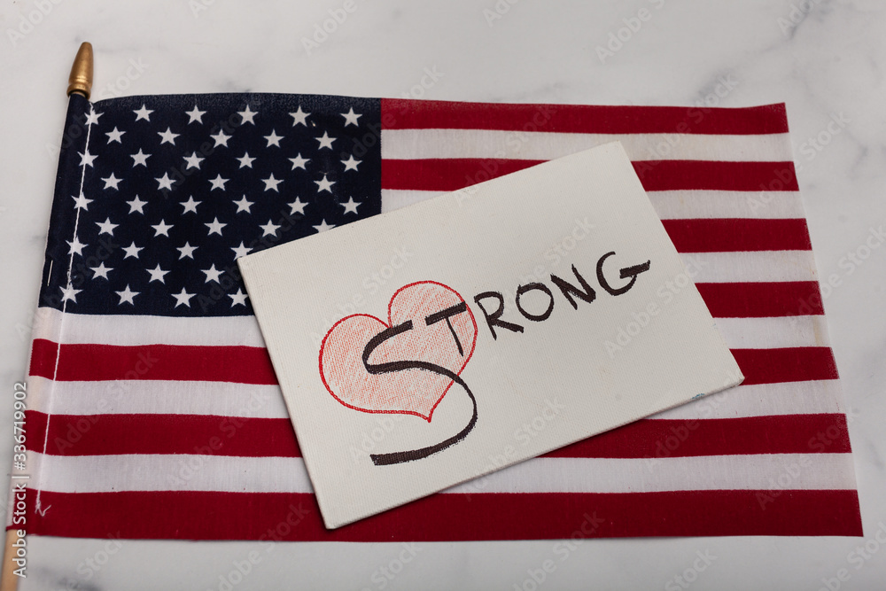 Fototapeta premium Red heart with the word strong painted in black on white canvas with an American flag laying in the background 