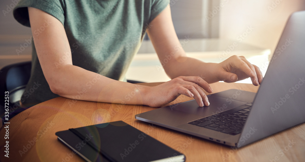 Naklejka premium Young woman in green t-shirt sitting at the wooden table at home with laptop and notebook, working or shopping online. Close-up shot with only arms seen