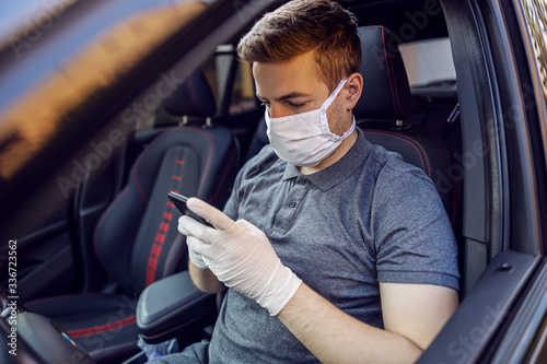 Man with protective mask and gloves driving a car holding mobile phone smartphone. Infection prevention and control of epidemic. World pandemic. Stay safe.